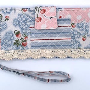 May include: A quilted wallet with a wrist strap. The wallet features a patchwork design with floral and cherry patterns in blue, pink, and white. A cream-colored lace trim adorns the bottom edge. The wrist strap is patterned in blue and red.