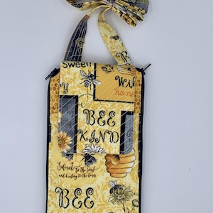 May include: A yellow and black quilted fabric phone case with a handle. The case features bee and honeycomb designs, with the words "BEE KIND" and "Sweet to the soul and healing to the bones" printed on it. The handle is made of a patterned fabric.