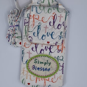 May include: White fabric phone bag with a handle and a small flap. The bag is covered in colorful script that reads "hope", "love", and "blessed", with cross and heart accents. The words "Simply Blessed" are embroidered on an oval patch.