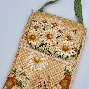 May include: A quilted crossbody bag with a daisy floral pattern in cream and tan. The bag features a green polka dot strap and the text "All Things Grow With Love". The bag has a zippered closure and a front pocket.