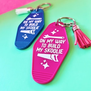May include: Two motel-style keychains in blue and hot pink, each featuring a white tassel and a metal ring. The text ON MY WAY TO BUILD MY SKOOLIE is printed in white, accompanied by graphic icons of a wrench, a hand saw, and four-pointed stars.