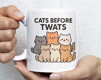 May include: White ceramic mug featuring the text "CATS BEFORE TWATS" above a group of six cartoon cats in various colors. The cats have smiling faces and are arranged in a row. The mug has a white handle.