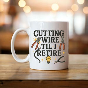 May include: White ceramic mug with the text "CUTTING WIRE TIL I RETIRE" in black. The mug features illustrations of wire cutters, a lightbulb, and stars. The mug is on a wooden surface.