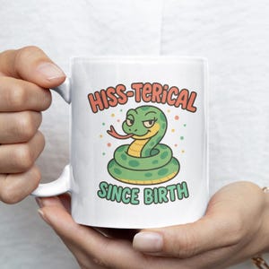 Puede incluir: Taza de cerámica blanca con un diseño de serpiente de dibujos animados y el texto "HISS-TERICAL SINCE BIRTH". La serpiente es verde con detalles amarillos y una lengua roja. El texto está en una fuente juguetona.