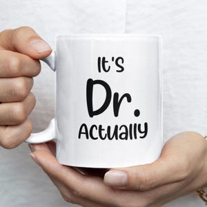 Könnte beinhalten: Weiße Keramik-Tasse mit dem Text "It's Dr. Actually" in Schwarz. Die Tasse hat einen Standardgriff und wird von einer Person gehalten.