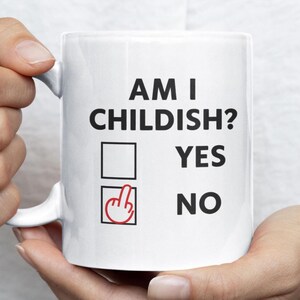 Könnte beinhalten: Weiße Keramik-Tasse mit dem Text "AM I CHILDISH?" in Schwarz. Darunter zwei Kästchen mit den Optionen "YES" und "NO". Das Kästchen "NO" hat eine rote Handgeste. Ein humorvoller Artikel.