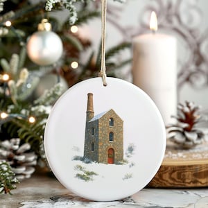 Könnte beinhalten: Runder, weißer Weihnachtsschmuck mit einer detaillierten Illustration eines Steingebäudes mit hohem Schornstein in einer verschneiten Landschaft. Das Gebäude hat eine rote Tür und gewölbte Fenster. Ein brauner Faden ist zum Aufhängen befestigt.