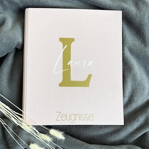 Könnte beinhalten: Ein hellrosa Hardcover-Buch mit dem Namen "Laura" und dem goldenen Buchstaben "L". Das Wort "Zeugnisse" ist unten in Gold aufgedruckt. Das Buch liegt auf einer grauen Decke mit weißen Trockenblumen.