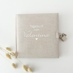 Könnte beinhalten: Ein beigefarbenes Leinen-Tagebuch mit Schloss und Schlüssel. Das Tagebuch trägt den Text "Tagebuch von Valentina" und ein kleines Herz auf der Vorderseite.