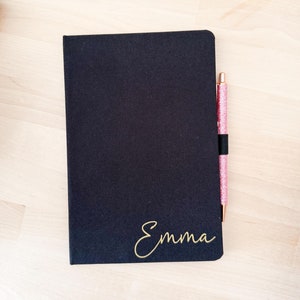 Op de afbeelding: Een zwart notitieboek met de naam "Emma" in gouden letters. Een roze glitterpen is gestoken in het elastiek op de omslag.