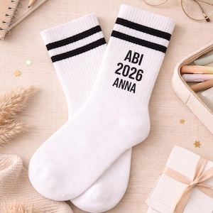 Könnte beinhalten: Ein Paar weiße Socken mit schwarzen Streifen am oberen Rand. Auf einer Socke steht in schwarzer Schrift "ABI 2026 ANNA". Die Socken liegen auf einer hellen Holzoberfläche, zusammen mit einem Notizbuch, Bleistift und anderen Gegenständen.