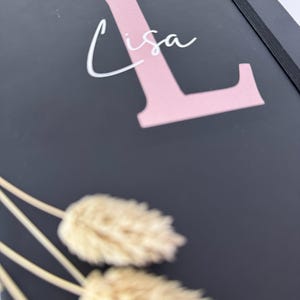 May include: A black notebook with a pink letter L and the name "Lisa" written in white. The notebook has a black elastic band closure and is lying on a surface with dried pampas grass.