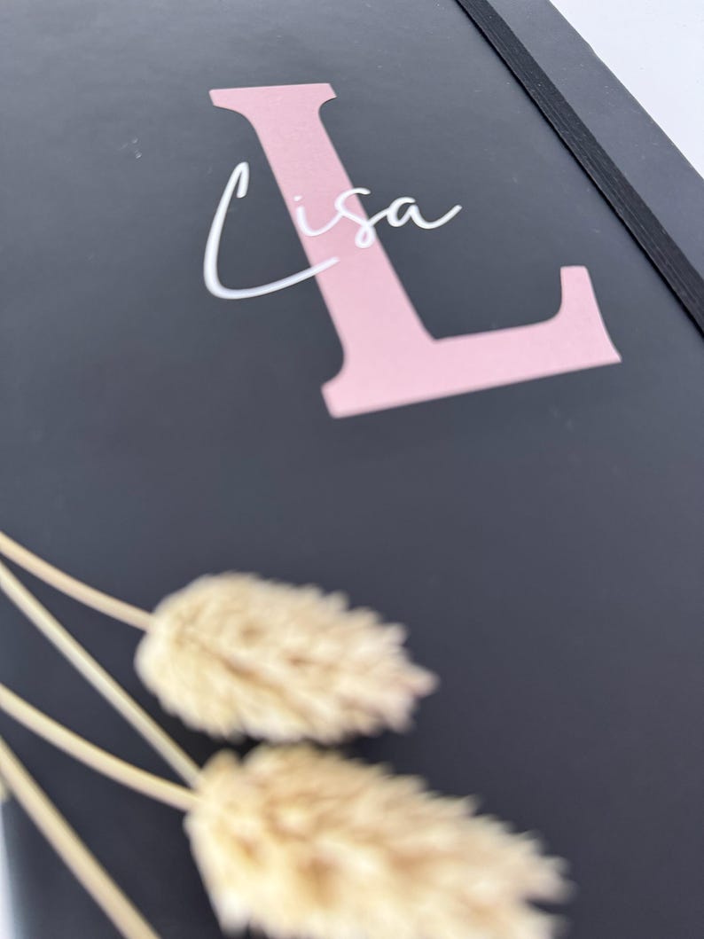 May include: A black notebook with a pink letter L and the name "Lisa" written in white. The notebook has a black elastic band closure and is lying on a surface with dried pampas grass.