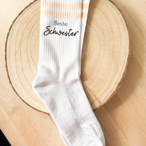 Könnte beinhalten: Weiße Socken mit zwei pfirsichfarbenen Streifen und dem Text "Beste Schwester" in Schwarz.