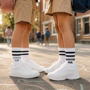 Könnte beinhalten: Zwei Beinpaare mit weißen Turnschuhen und weißen Socken mit schwarzen Streifen. Auf einem Paar Socken steht "SCHULKIND LIA" und auf dem anderen "SCHULKIND PAUL". Die Personen tragen khakifarbene Shorts und Rucksäcke.