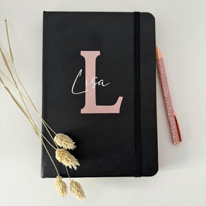 May include: A black notebook with a rose gold elastic closure and a rose gold letter "L" with the name "Lisa" written in white script.