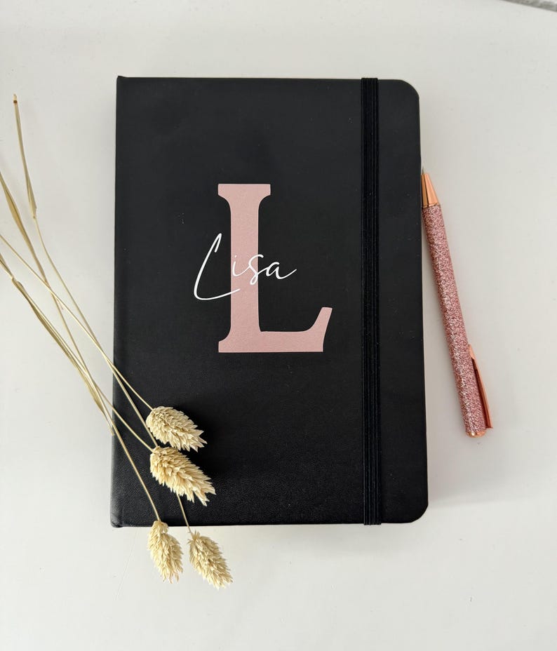 May include: A black notebook with a rose gold elastic closure and a rose gold letter "L" with the name "Lisa" written in white script.