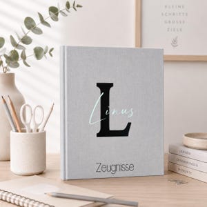 Könnte beinhalten: Ein hellgraues Notizbuch mit dem schwarzen Buchstaben "L" und dem Namen "Linus" in Schreibschrift. Das Wort "Zeugnisse" ist unten aufgedruckt. Das Notizbuch liegt auf einer Holzoberfläche.