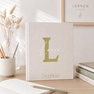 Könnte beinhalten: Ein beiges Notizbuch mit dem Namen "Laura" und dem Anfangsbuchstaben "L" in goldfarbenen Buchstaben. Das Wort "Zeugnisse" ist unten aufgedruckt. Stifte und eine Vase mit Trockenblumen sind im Hintergrund.