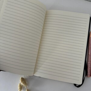 May include: An open lined notebook with a black cover and a black pen lying on a white surface.
