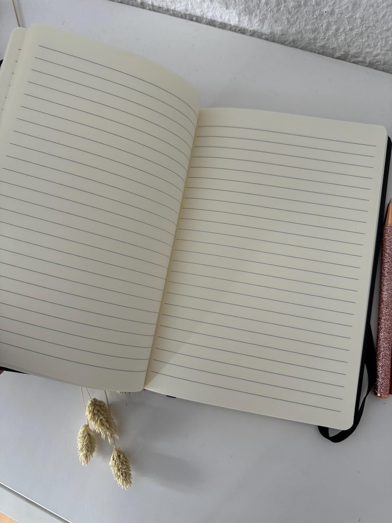 May include: An open lined notebook with a black cover and a black pen lying on a white surface.