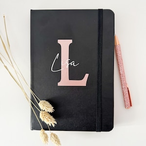 Könnte beinhalten: Schwarzes Notizbuch mit einem roségoldenen Buchstaben L und dem Namen Lisa in weißer Schrift. Das Notizbuch hat einen schwarzen Gummibandverschluss und liegt auf einer weißen Oberfläche.
