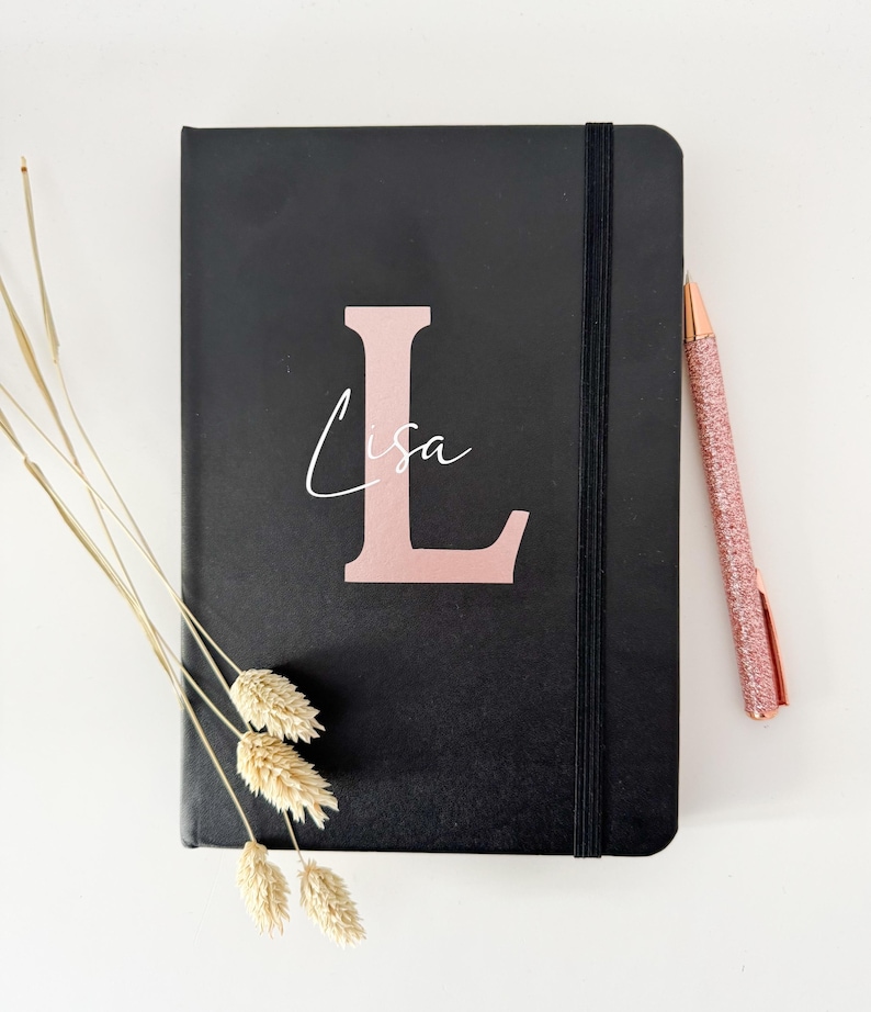May include: Black notebook with a rose gold letter L and the name Lisa written in white script. The notebook has a black elastic band closure and is lying on a white surface.