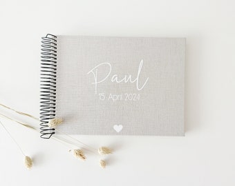 Op de afbeelding: Een grijs linnen gebonden spiraalgebonden fotoalbum met de naam "Paul" en de datum "15 april 2024" in wit gedrukt. Een klein wit hartje is onder de datum gedrukt.