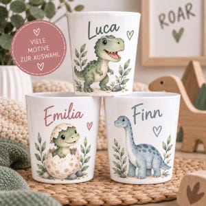 Op de afbeelding: Drie witte cilindrische bekers met kleurrijke dinosaurusillustraties en gepersonaliseerde namen. De bovenste beker toont een groene T-Rex met de naam Luca. De linkeronderbeker toont een babydinosaurus in een ei met de naam Emilia. De rechteronderbeker toont een blauwe langnekdinosaurus met de naam Finn.