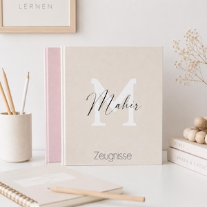 Op de afbeelding: Een beige gepersonaliseerd notitieboek met de naam "Mahir" in script en een grote witte "M". Het woord "Zeugnisse" staat eronder gedrukt. Een roze notitieboek staat erachter, met potloden en een notitieblok op de achtergrond.