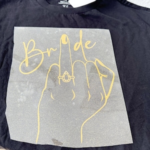 Könnte beinhalten: Schwarzes T-Shirt mit einem goldenen Glitzer-Bügelbild einer Hand mit einem Ring am Mittelfinger und dem Wort "Bride" darüber.
