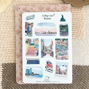 May include: A white sticker sheet featuring multiple watercolor-style illustrations of Busan, South Korea. The text A Day in Busan is printed at the top. The images include cityscapes, a bridge, a cable car, street scenes, and a blue street sign.