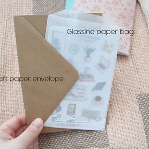 May include: A brown kraft paper envelope containing a translucent glassine paper bag. Inside the bag is a sticker sheet featuring various illustrations including a potted plant, a laptop, books, and text that reads A-Nook and anook5.com.