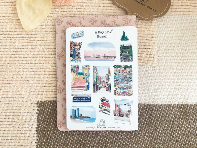 May include: A white sticker sheet featuring multiple watercolor-style illustrations of Busan, South Korea. The text A Day in Busan is printed at the top. The images include cityscapes, a bridge, a cable car, street scenes, and a blue street sign.