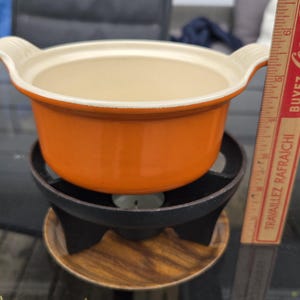 May include: Orange ceramic fondue pot with white interior and handles, set on a black metal stand with a wooden base. A Coca-Cola ruler is next to the pot. The pot is approximately 7.6 cm tall.