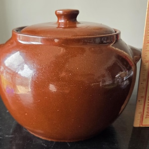 May include: A brown ceramic casserole dish with a lid and two side handles. The dish has a glossy finish and a small knob on top of the lid. A wooden ruler with Coca-Cola branding is next to the dish.