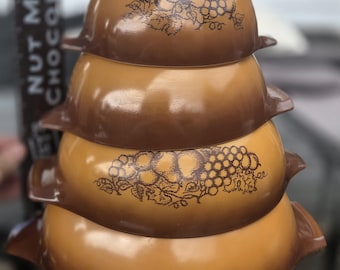 May include: A stack of four graduated brown glass mixing bowls with side handles. The top and third bowls feature a dark brown printed graphic of grapes and leaves. A black ruler with the text NUT MILK CHOCOLATE is visible on the left side.
