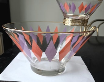 May include: A vintage glass serving set featuring a large bowl and a smaller, elevated bowl. Both bowls are clear with a gold rim and decorated with orange and purple diamond shapes. The set is likely from the mid-century era.