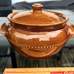Peut inclure: Une cocotte en céramique émaillée marron avec un couvercle assorti, dotée de deux poignées latérales et d'une texture perlée décorative autour du centre et du couvercle. Une règle en bois jaune est placée à la base, indiquant 15 cm de largeur.
