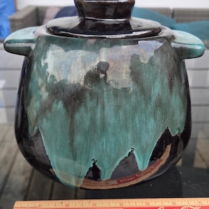 May include: A ceramic lidded jar with two side handles, featuring a glossy black base and a flowing teal glaze drip pattern. A wooden ruler placed in front shows the jar is approximately 15 cm wide at the base and 18 cm wide at the handles.