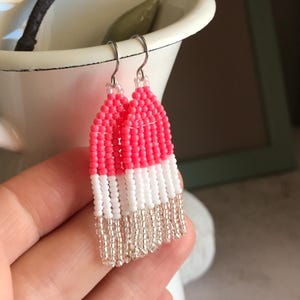 Puede incluir: Pendientes hechos a mano con cuentas rosas, blancas y plateadas. Los pendientes tienen un diseño rectangular con flecos de cuentas. Se exhiben frente a un jarrón blanco con un arreglo floral.