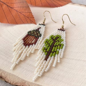 Pode incluir: Brincos pendurados com contas com tema de outono. Um brinco apresenta um design de bolota marrom e vermelha, o outro uma árvore verde e marrom. Ambos têm franjas de contas brancas e ganchos dourados.