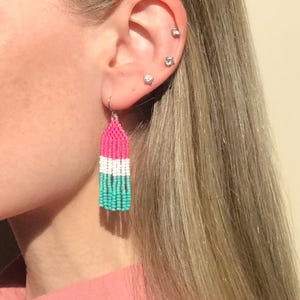 Puede incluir: Primer plano de un pendiente de cuentas con una combinación de colores rosa, blanco y turquesa. El pendiente cuelga de un gancho plateado. El pendiente lo lleva una persona con pelo rubio y una camisa rosa.