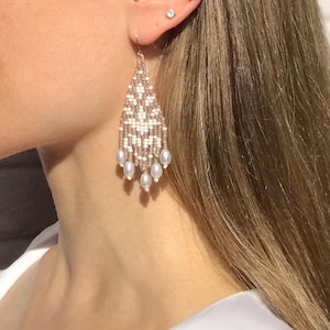 May include: A pair of long, beaded dangle earrings with a pink and white color scheme. The earrings feature a teardrop shape with a fringe of white pearls at the bottom.