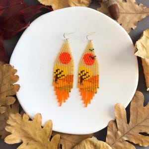 Puede incluir: Pendientes de cuentas con un diseño de paisaje otoñal. Los pendientes presentan una puesta de sol con un sol rojo, árboles amarillos y naranjas, y una franja de cuentas naranjas. Los pendientes están sobre un plato blanco.