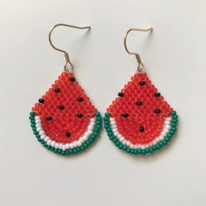 May include: A pair of watermelon-shaped earrings made with red, green, white, and black beads. The earrings are hanging from gold hooks.