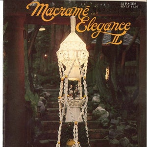 May include: The cover of a vintage macrame book, "Macrame Elegance II," features a detailed white macrame hanging lamp. The lamp has multiple tiers, tassels, and decorative elements. The background shows a blurred outdoor scene with stairs and foliage.
