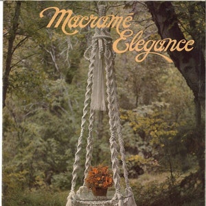 May include: A white macrame plant hanger with a glass top and a fringe bottom. The hanger is hanging in a forest setting with green trees and brown ground. The title "Macrame Elegance" is written in cursive on the cover of the book.