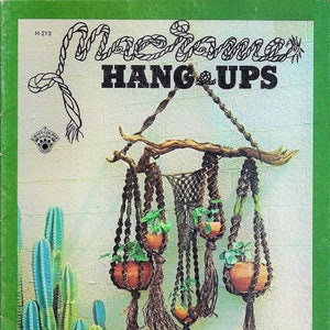 May include: A vintage book cover titled "Macrame Hang Ups" featuring a macrame plant hanger with terracotta pots and green plants. The cover has a green border and a cactus in the background.