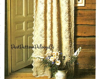 May include: A crocheted curtain with a geometric pattern hangs in a rustic wooden room. The curtain is cream colored and has a scalloped edge. A vase of flowers sits on a wooden surface in front of the curtain.